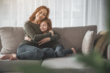 My dearest. Full length portrait of mother and her child hugging each other and sitting on couch at home. They are smiling, calm and happy