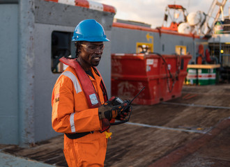 Seaman AB or Bosun on deck of offshore vessel or ship , wearing PPE personal protective equipment - helmet, coverall, lifejacket, goggles. He holds VHF walkie-talkie radio in hands. Cargo operations