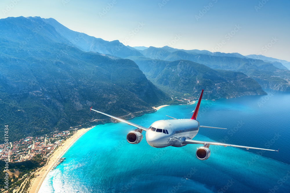 Airplane is flying over amazing mountains with forest and sea at sunrise in summer. Landscape with white passenger airplane, sky, islands and blue water. Passenger aircraft. Travel and resort. Tourism - obrazy, fototapety, plakaty 