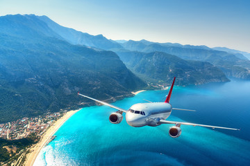 Airplane is flying over amazing mountains with forest and sea at sunrise in summer. Landscape with white passenger airplane, sky, islands and blue water. Passenger aircraft. Travel and resort. Tourism