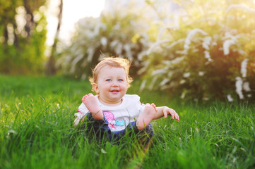  A funny baby girl is playing and smiling at the bright green grass in the park in the summer.