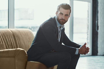 Young bearded stylish businessman leader indoors at office sitting looking aside holding sunglasses thoughtful