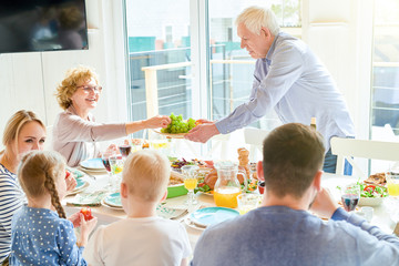 Portrait of happy two generation  family enjoying dinner together sitting round festive table handing delicious dishes and laughing during  holiday  celebration in modern sunlit apartment, copy space