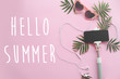 Hello Summer text on stylish pink sunglasses, phone on selfie stick, headphones, and green palm leaves on pink background. stylish summer vacation flat lay. hello holidays