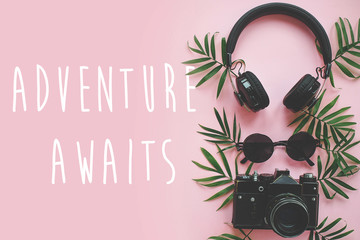 Wall Mural - The Adventure Awaits text on stylish photo camera with green palm leaves, sunglasses and headphones on trendy pink paper. modern summer vacation image, stylish flat lay