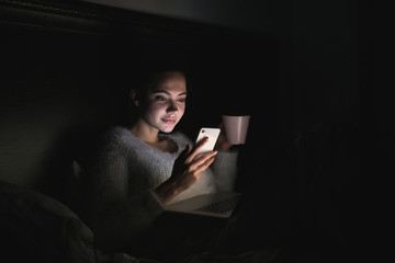 a cute young girl sits on a bed in the dark at night, watching a movie on a laptop, drinking tea and holding a smartphone in her hand