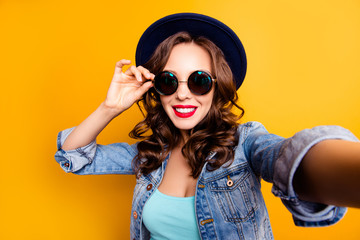 Wall Mural - Portrait of trendy pretty tourist with beaming smile shooting self portrait on front camera holding eyelet of glasses on face wearing jeans jacket isolated on yellow background, travel tourism concept