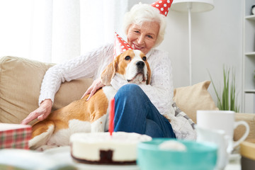 Wall Mural - Portrait of happy senior woman hugging dog sitting on couch while celebrating birthday at home with festive dinner table with cake in foreground, copy space