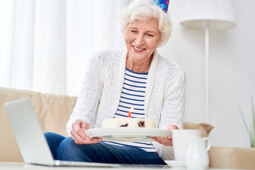 Wall Mural - Portrait of happy senior woman showing cake to camera while celebrating birthday with her family via video chat