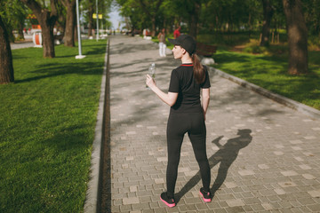 Back view Young athletic beautiful brunette girl in black uniform and cap holding bottle, with water during training before running standing in city park outdoors. Fitness, healthy lifestyle concept.