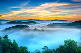 Fototapeta Na ścianę - Sunrise over hillside as the sun rising from horizon reflect light bright yellow sky. Below cloudy mist covered valleys flooded pine forests create impressive beauty highlands in morning.