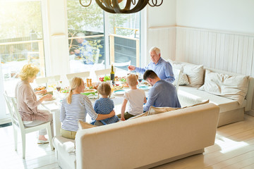 Back view of happy two generation family with two kids enjoying dinner together sitting round festive table during  holiday  celebration in modern sunlit apartment, copy space