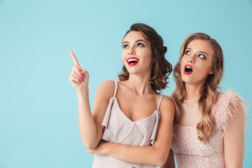 Poster - Two beautiful girls 20s wearing party outfit pointing index fingers upward at copyspace with excited faces, isolated over blue background