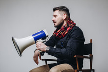 Young Attractive Movie Director Holding A Megaphone. Film Director Talking On A Loudspeaker While Sitting On A Chair Isolated On Grey Background. Side View