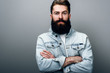 © iuricazac - Brutal handsome fashionable young European male with thick beard wearing trendy denim jacket, cross hands, staring at camera with serious and cheerful look. Barber man posing against gray studio wall.