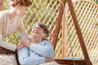An elderly female pensioner with disabilities sitting on a patio swing during rehabilitation camp. Professional caretaker standing next to the woman. Wooden fence behind.