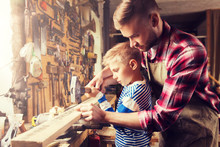 Family, Carpentry, Woodwork And People Concept - Father And Little Son With Hammer Hammering Nail Into Wood Plank At Workshop