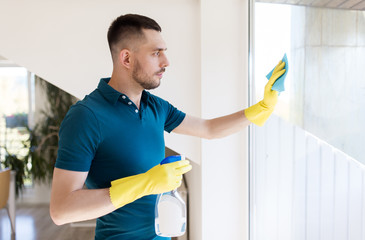 Wall Mural - household and people concept - man in rubber gloves cleaning window with rag and spray cleaner at home