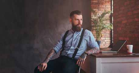 Handsome tattooed hipster in a shirt and suspenders sitting at the desk with a computer, looking out the window in an office with loft interior.