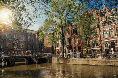 Zdjęcie XXL Most z żelazną balustradą na wysadzanym drzewami kanale, starych budynkach i słonecznym niebie w Amsterdamie. Miasto słynie z ogromnej aktywności kulturalnej, pięknych kanałów i mostów. Północnej Holandii.