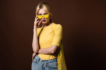 stylish blonde girl with string bag wearing yellow sunglasses and looking at camera isolated on brown