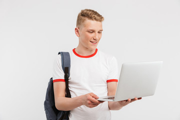 Canvas Print - Image of teenage guy 16-18 years old wearing casual clothing and backpack holding laptop and looking at screen, isolated over white background