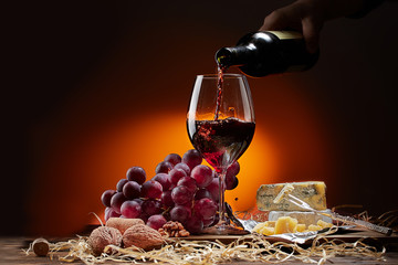 Wine flows from the bottle into the glass. On the table are different varieties of cheese and grapes. Dark background.