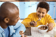 © zinkevych - More delicious together. Joyful pre-teen boy sitting at the table next to his father and smiling at his father while eating cereals together with him