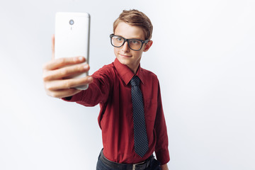 Portrait of a positive and emotional schoolboy, takes a selfie on your smartphone, white background, glasses, red shirt, business theme, promotional,