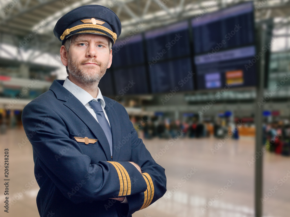 Pilot in der Uniform mit goldenen Streifen und Kappe kreuzte seine Arme beim Stehen im Flughafen - obrazy, fototapety, plakaty 