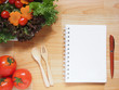 Blank spiral notebook for recipe or diet plan with pen, spoon, fork, tomatoes and salad on wooden table. Healthy lifestyle concept. Copy space.