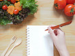 Woman’s hand writing recipe or diet plan on blank spiral notebook with salad, tomatoes, spoon and fork on wooden table. Diet control or Healthy lifestyle concept. Space for your text.