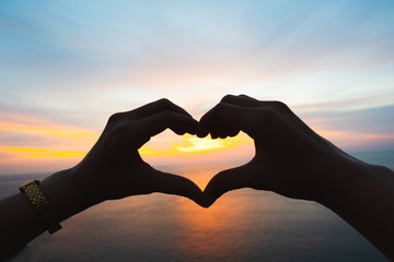 Couple hands in heart shape against sunlight in nature sunset sky and summer sea.