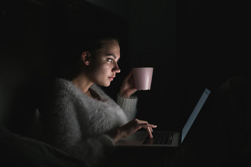 cute young freelancer girl sitting in bed late at night, working on laptop and drinking tea