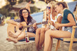 © BalanceFormCreative - Group of young female friends sitting on beach on sunbeds,singing and playing guitar.