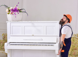 Wall Mural - Courier delivers furniture, move out, relocation. Man with beard worker in helmet and overalls lifts up, efforts to move piano, white background. Loader moves piano instrument. Heavy loads concept.