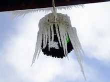 Big Icicles Hanging On An Electric Lantern With Green And Clear Glass And A Light Bulb Inside. Karlova Studanka, Jeseniky, The Czech Republic. View Against Blue Sky. View From The Bottom