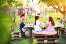 Adorable Children Are Having A Picnic Meal At The Table Under The Tree While Thier Parents Are Grilling In Backyard.