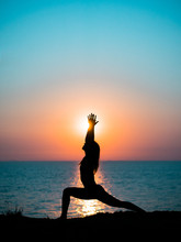 Young Slim Girl Practicing Yoga On Mountain Against Ocean Or Sea At Sunrise Time. Silhouette Of Woman In Rays Of Awesome Sunset.