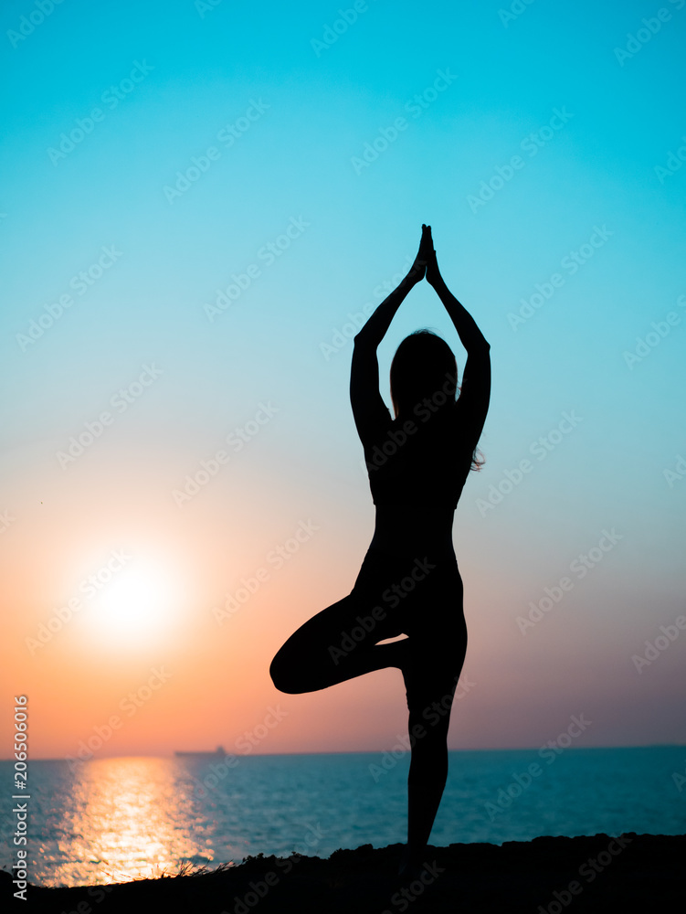 Young slim girl practicing yoga on mountain against ocean or sea at sunrise time. Silhouette of woman in rays of awesome sunset. - obrazy, fototapety, plakaty 