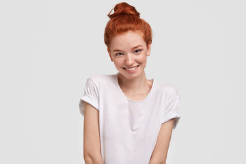 Wall Mural - Portrait of happy red haired female being in good mood, wears casual clothing, has shy expression, recieves compliment, stands against white background. Isolated shot. People and skin care concept