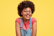Positive glad African American female has broad smile, wears spectacles, dressed in denim overalls, poses against yellow background. Pretty delighted woman poses in studio alone. Emotions concept