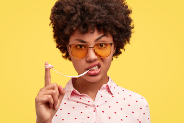 Photo of good looking young African American female has angry expression, stretches bubble gum, feels bored and annoyed as waits friend for long time, wears formal blouse and trendy yellow sunglasses