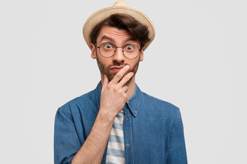 Wall Mural - Portrait of shocked man with stubble, keeps hand on chin, looks with stunned expression at camera. Surprised male tourist in glasses, wears straw hat and denim shirt, poses against white background