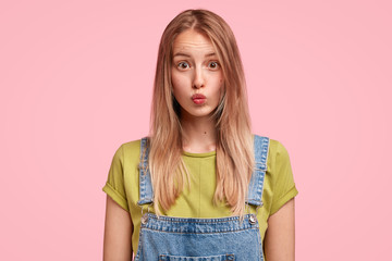 Wall Mural - Horizontal portrait of pleasant looking female keeps lips round, has long hair, expresses bewilderment as recieves proposal from friend to date, isolated over pink background. Teenager makes grimace