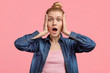 Emotive stupefied young European female keeps mouth widely opened as sees something incredible, keeps hands on head, stares at camera, poses against pink background. People and shock concept
