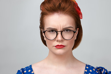 Wall Mural - Close up shot of attractive young Caucasian female teacher wearing dotted dress, red lipstick and stylish eyeglasses frowning, looking at camera with strict expression, annoyed with noisy pupils