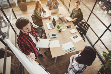 Top view pleased man telling with happy lady while standing near board. Outgoing colleagues listening them while sitting at desk. Cheerful comrades at job concept