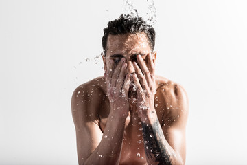 Portrait of young shirtless man with tattooed hand is standing against light wall and washing his face in the morning. Drops of water around him. Skin care and male beauty concept