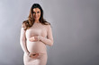 © Spectral-Design - A young happy pregnant woman in an elegant dress standing with hands on her belly in front of a grey background in a studio.